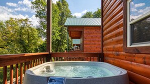 A hot tub on a wooden deck with a view of a cabin and trees.