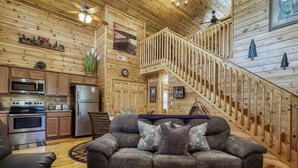 A cozy cabin interior with a wooden interior, a kitchen area, a sofa with cushions, and a staircase.