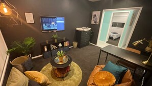 A modern living room with a dark wall, a round coffee table, a TV, a bookshelf, and a bed in the background.