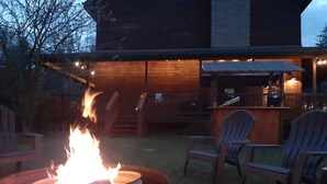 Una hoguera en un contenedor metálico circular, rodeada por dos sillas Adirondack, con una terraza de madera y un edificio al fondo.