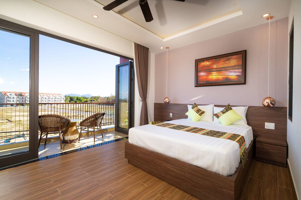 A modern hotel room with a large bed, a balcony with wicker chairs, and a city view.