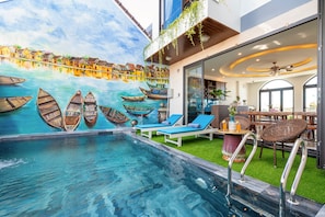 Una piscina con sillones de descanso, un mural de barcos y un área de restaurante con mesas y sillas.