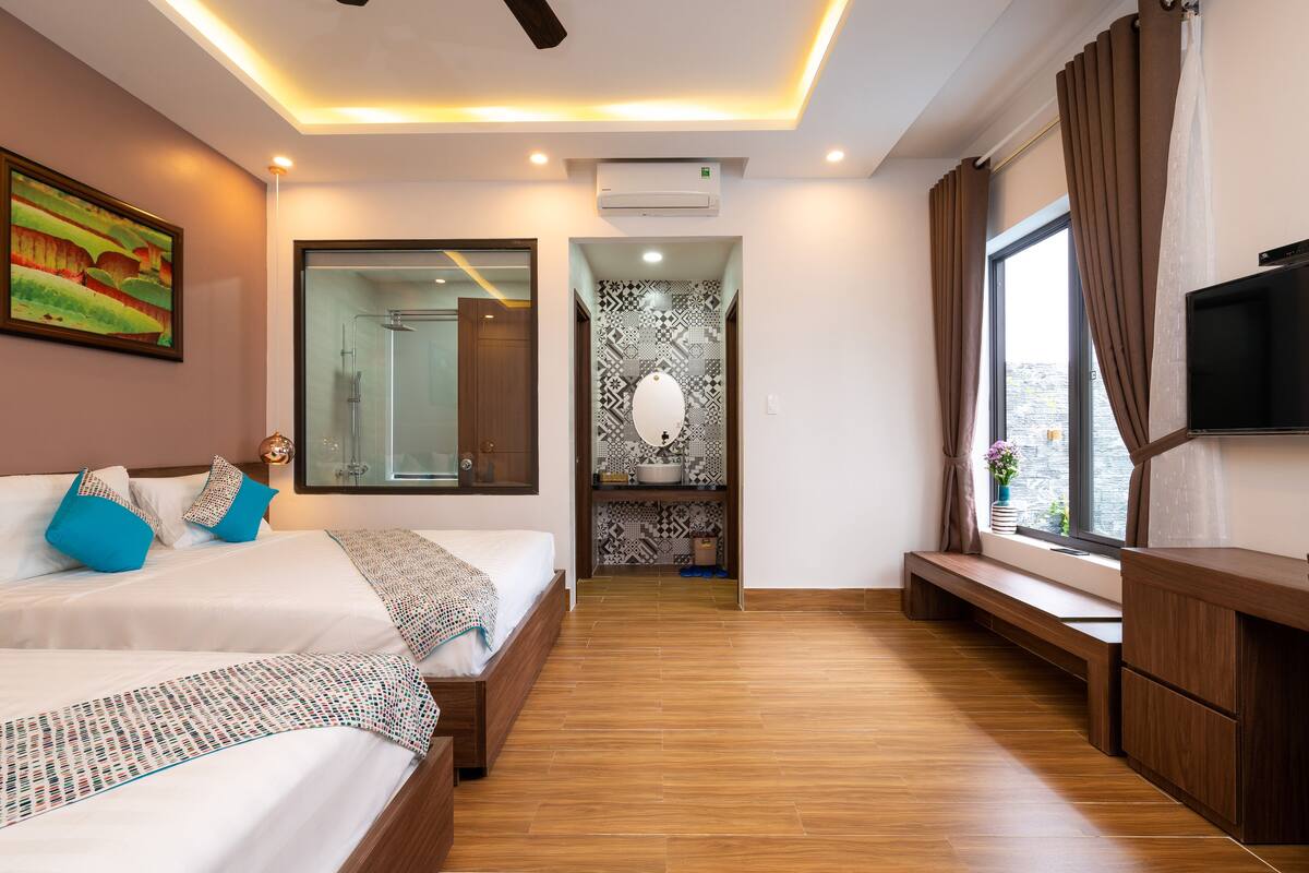 A modern hotel room with two beds, a TV, a large window, and a shower area.