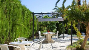 An outdoor patio with two round tables and white chairs, surrounded by greenery and a pergola.