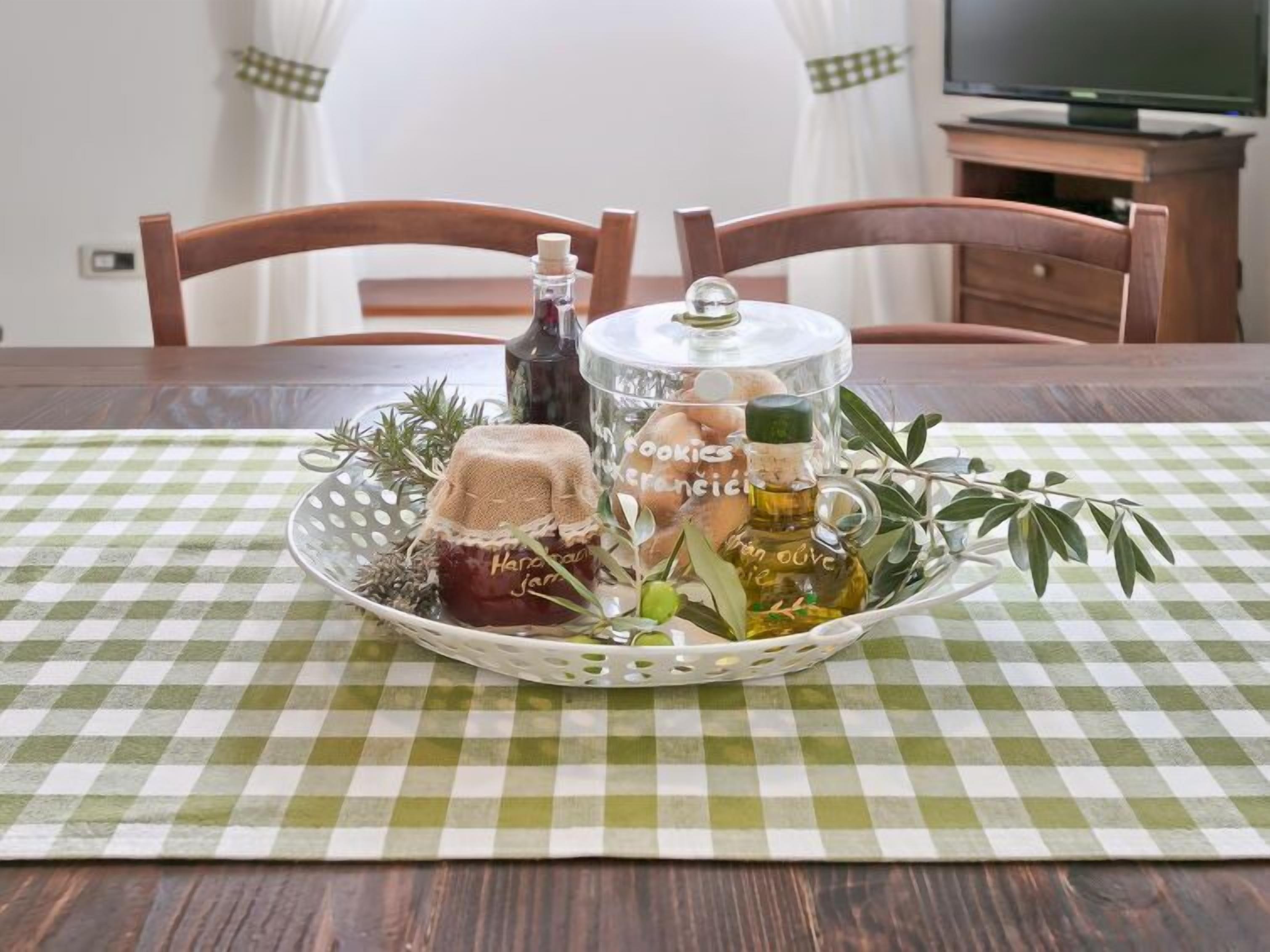 A dining table with a checkered tablecloth, a basket containing jars of homemade preserves, and a bottle of olive oil.