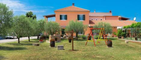 A two-story house with a red-tiled roof, a garden with trees and a swing set, and a clear blue sky.