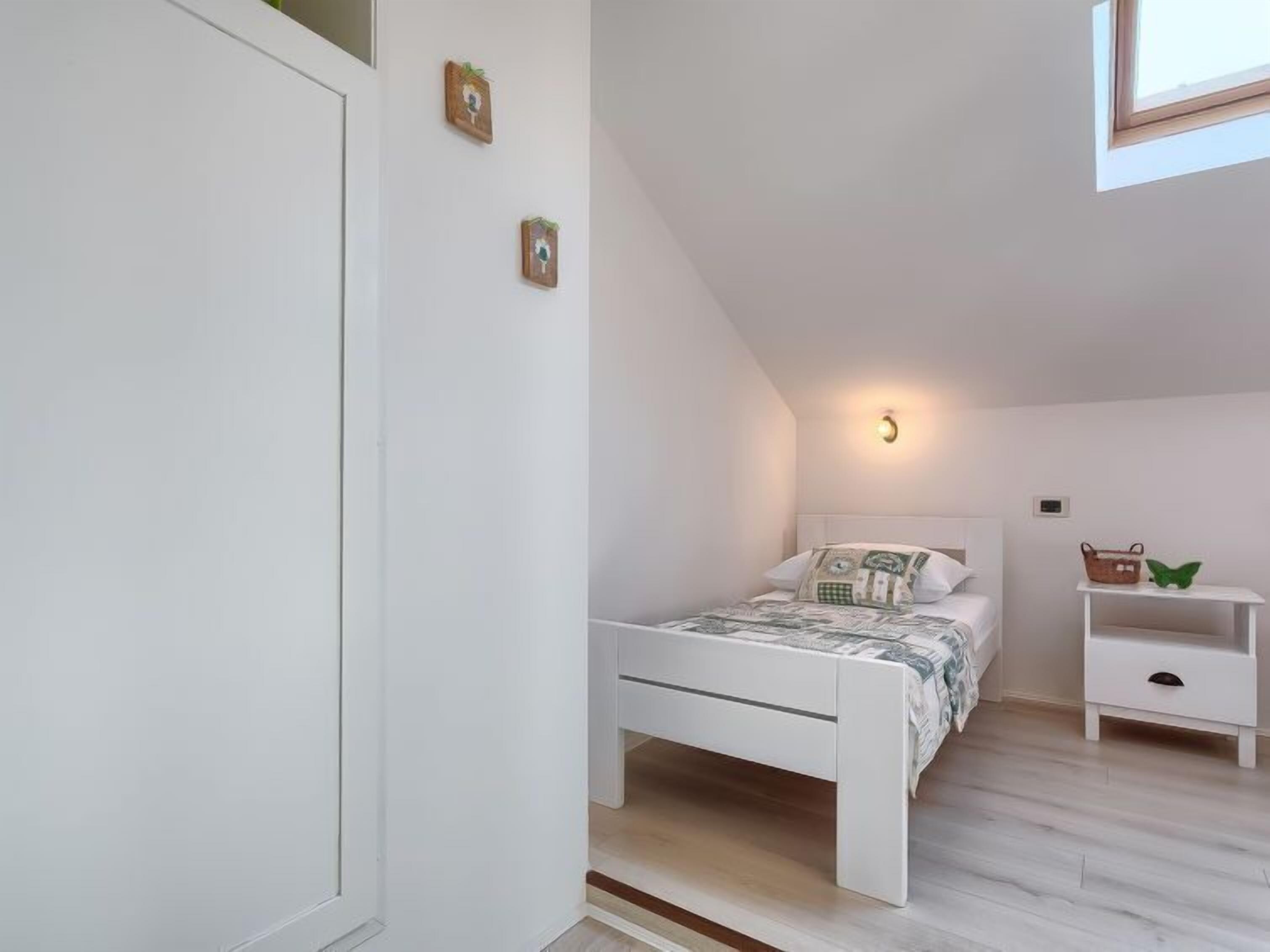 Ein Schlafzimmer mit einem weißen Bett, einem Nachttisch, einer Wandleuchte und einem kleinen Regal mit Korb und Dekoartikel.