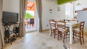 Zona pranzo con tavolo e sedie, un televisore su un mobile e vista all'esterno attraverso una finestra con tende.