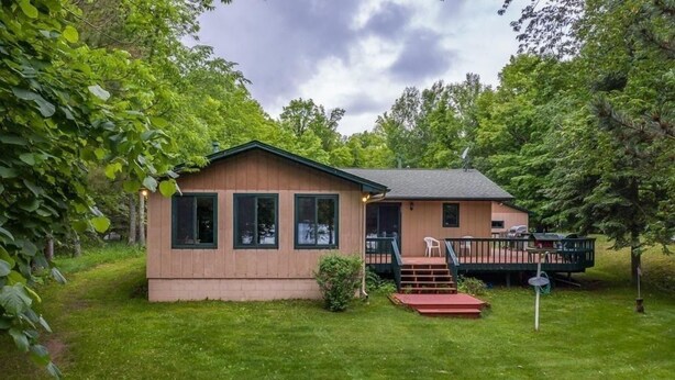 A single-story wooden cabin with a deck, surrounded by lush greenery.
