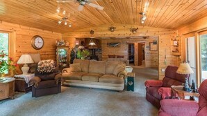 A cozy log cabin interior with a wooden ceiling, a large couch, a red armchair, and a dining area.