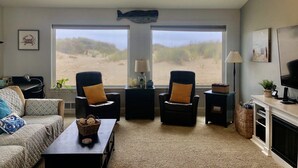 Ein Wohnzimmer mit großem Fenster mit Blick auf einen Strand, zwei Sessel, ein Sofa, ein Couchtisch und ein Kamin.