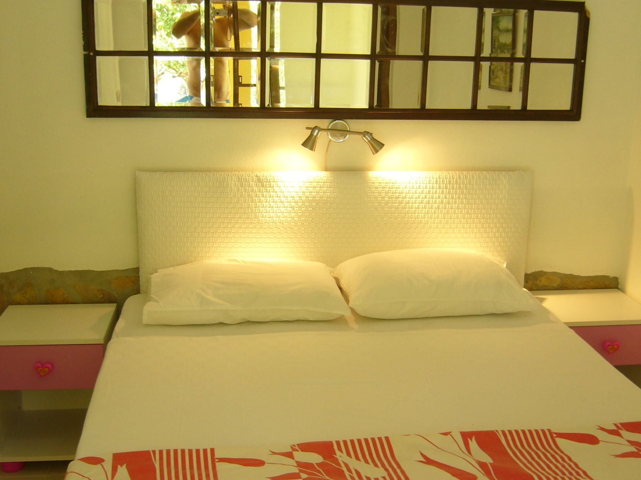 A neatly made bed with white linens and two pillows, a bedside table with a lamp, and a wall-mounted mirror above the bed.
