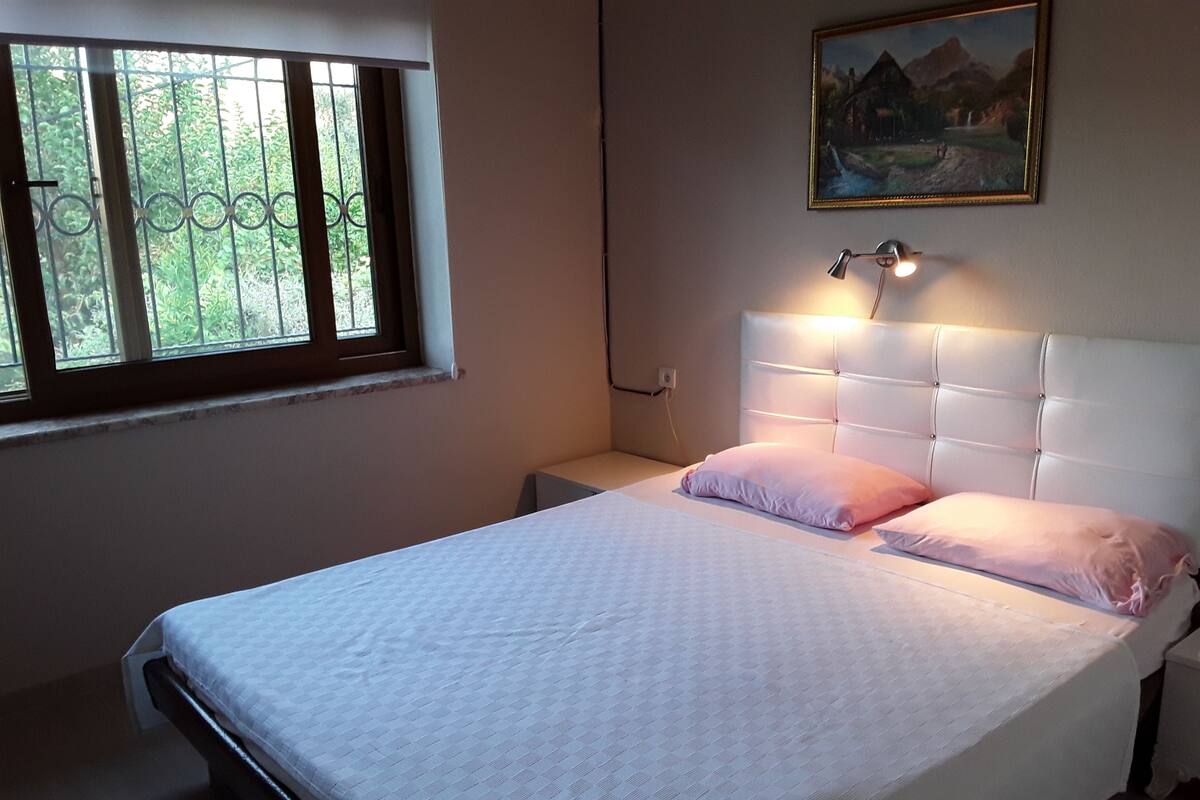 A bedroom with a bed, a nightstand, a window with bars, and a painting on the wall.