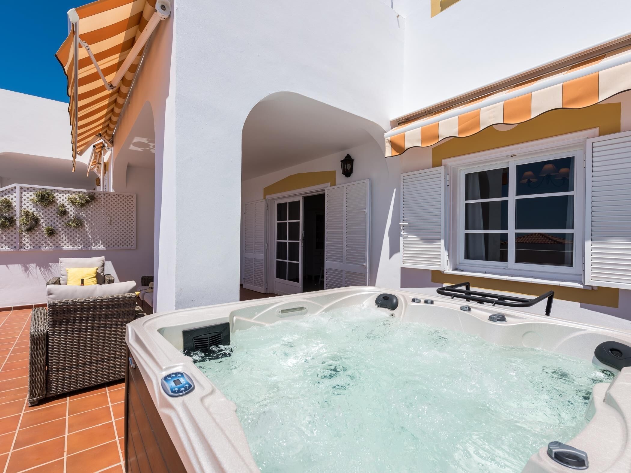 A hot tub on a terrace with a white wall, a wicker chair, and a door leading to an interior space.