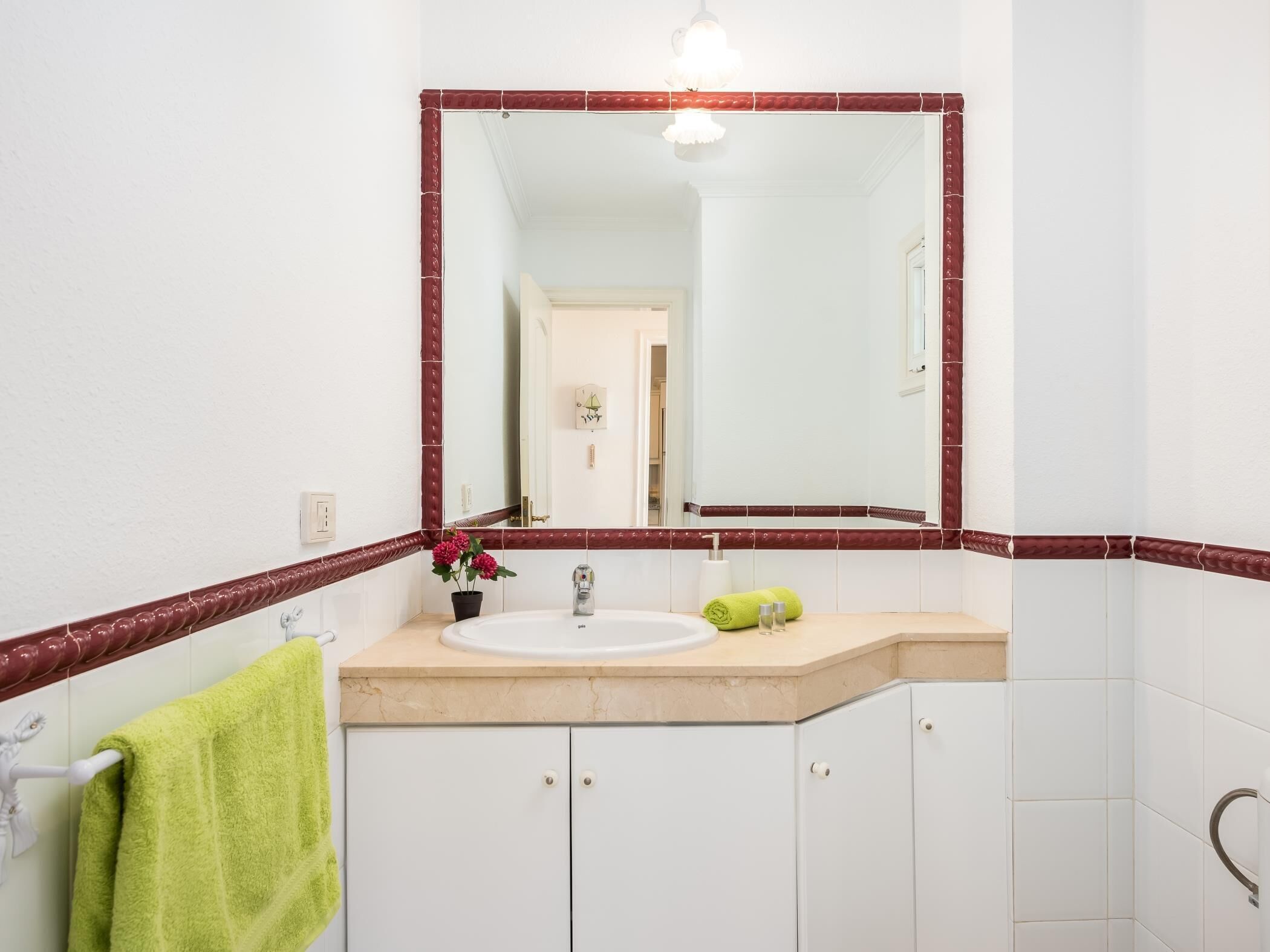 Een badkamer met een witte wasbak, een spiegel met rode rand en een groene handdoek op een rek.