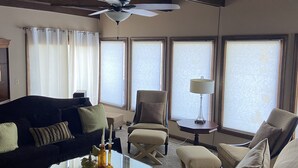 En stue med en mørk sofa, en beige lænestol og et glasbord. Der er en ventilator i loftet og store vinduer med skodder.