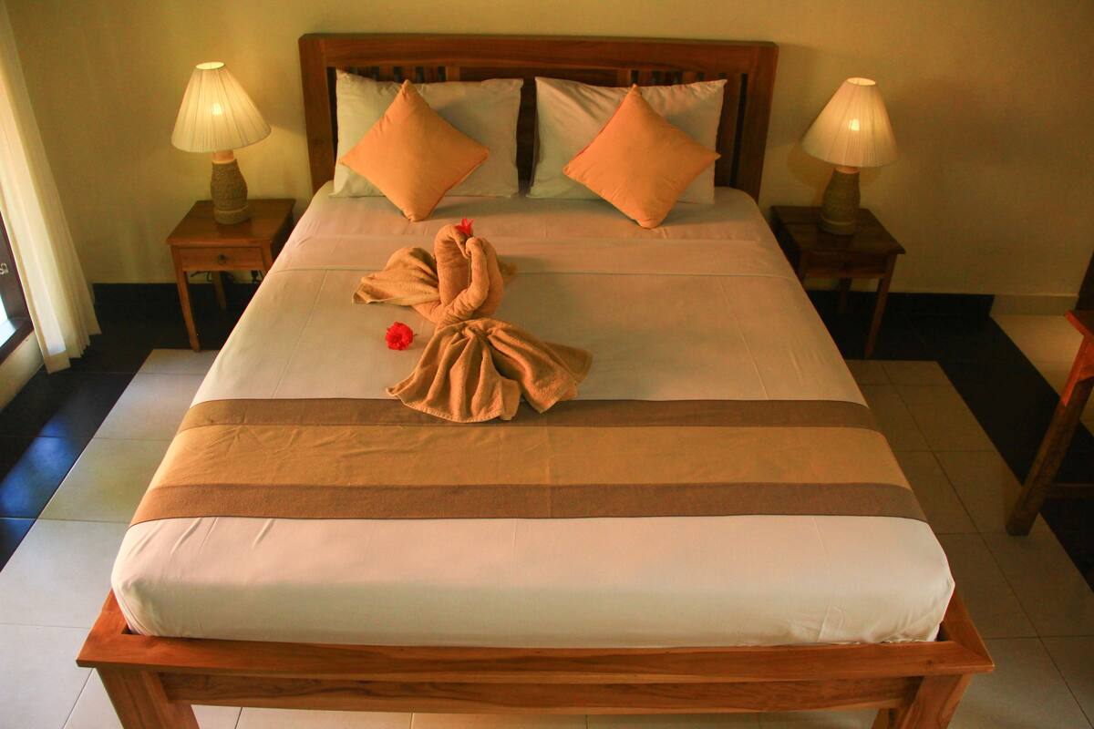 A neatly made bed with a brown and white striped bedspread, two matching pillows, and a towel arranged in a bow on the bed. There are two bedside tables with lamps on either side of the bed.