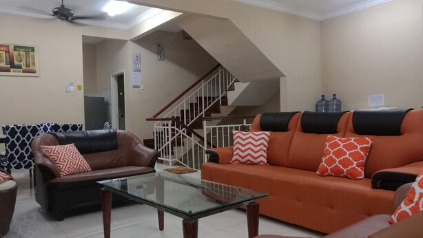 Una sala de estar con un sofá de cuero marrón, un sofá naranja y una mesa de centro de vidrio. Se observa una escalera con barandilla blanca que conduce a un piso superior.