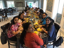 Un grupo de personas sentadas alrededor de una larga mesa de comedor en un restaurante con papel tapiz floral y ventanas grandes.