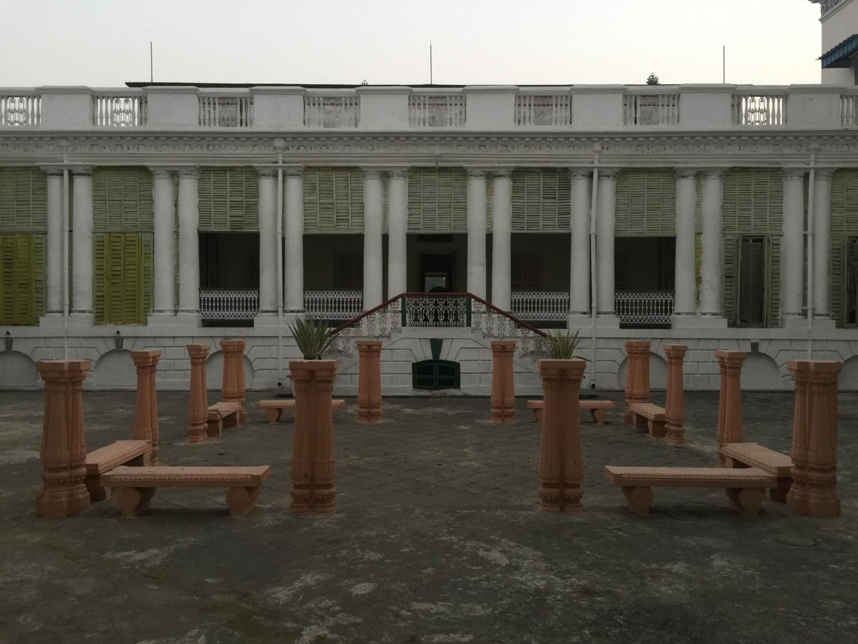 A courtyard with columns and benches, a staircase, and a building facade with windows and shutters.