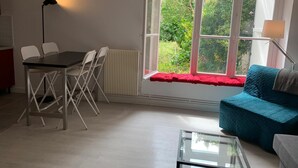 Ein Zimmer mit Esstisch, Stühlen, einer Couch, einem Glastisch und einem großen Fenster mit Blick ins Grüne.