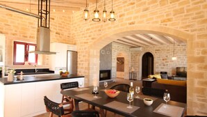 A modern kitchen with a stone wall, a dining area with a table set for two, and a cozy living space with a sofa and fireplace. - Villa Dionysos, luxury villa with a fantastic view of a large plot of land (Rethymnon)