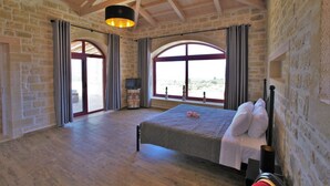 A spacious bedroom with a large bed, stone walls, wooden beams, and large windows offering a view of the outdoors. - Villa Dionysos, luxury villa with a fantastic view of a large plot of land (Rethymnon)
