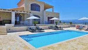 A poolside area with a pool, lounge chairs, umbrellas, and a building with a stone facade. - Villa Dionysos, luxury villa with a fantastic view of a large plot of land (Rethymnon)
