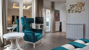 A modern living room with a teal armchair, a white coffee table, and a teal sofa. A television is mounted on the wall, and there is a radiator on the floor.