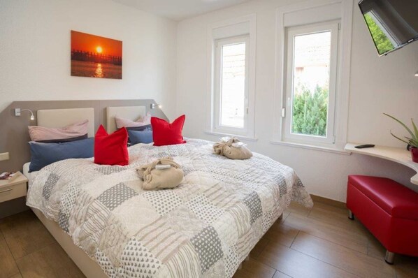 Un dormitorio con cama, almohadas, mesita de noche, ventana y televisor instalado en la pared.