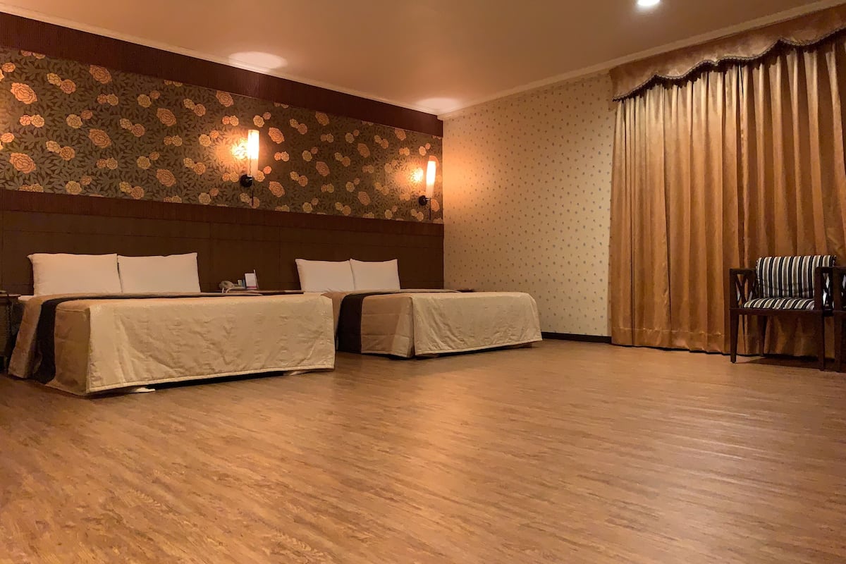 A hotel room with two beds, a wooden floor, a floral wallpaper design, and a large window with curtains.