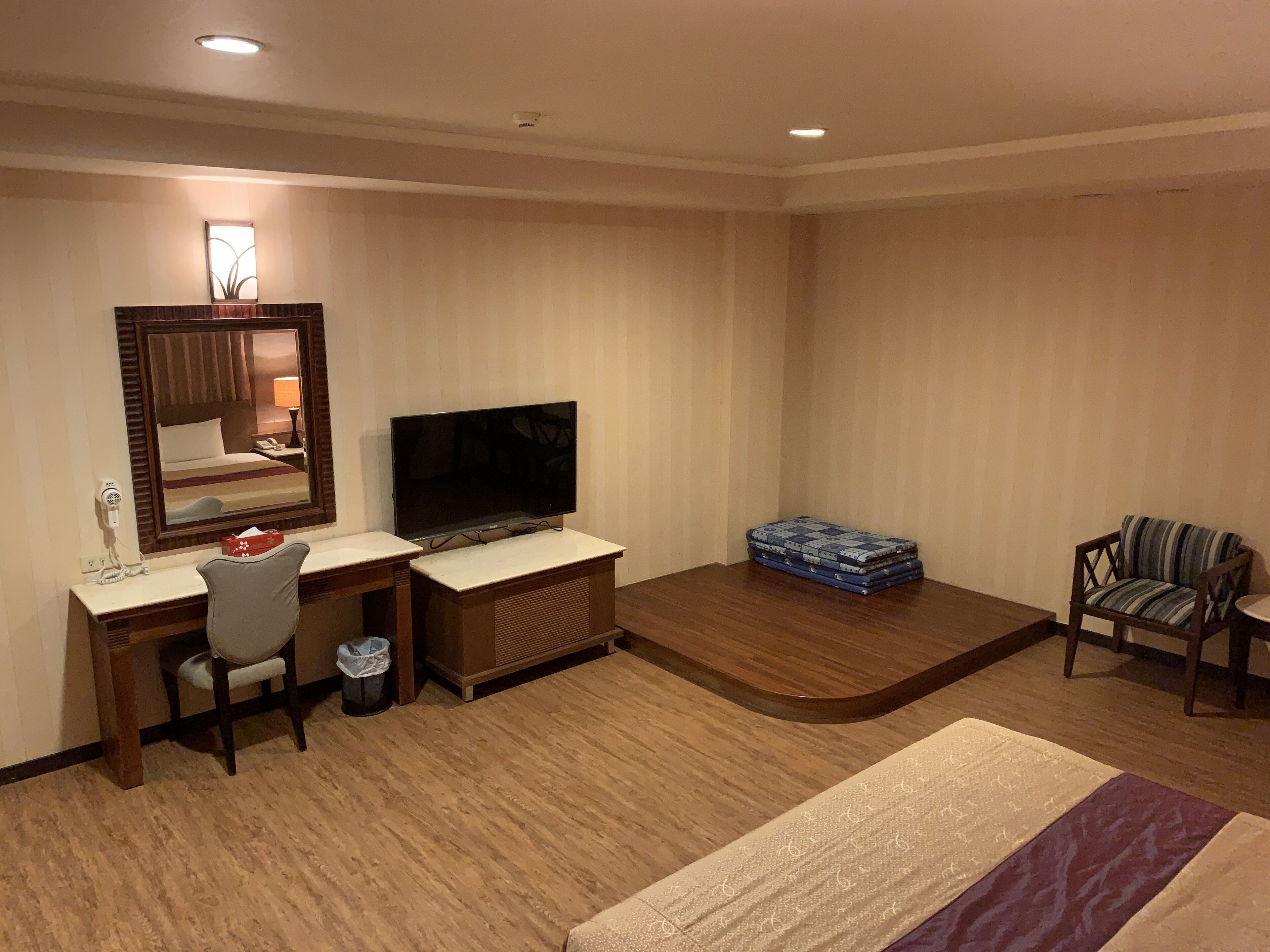 A hotel room with a desk, chair, TV, and a small seating area with a chair and a box of books.