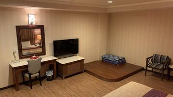 A hotel room with a desk, chair, TV, and a small seating area with a chair and a box of books.