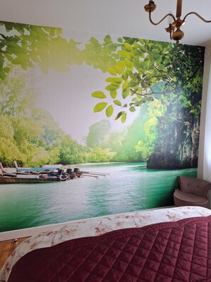 A bedroom with a large mural of a river scene, a bed with a floral bedspread, and a brown armchair.