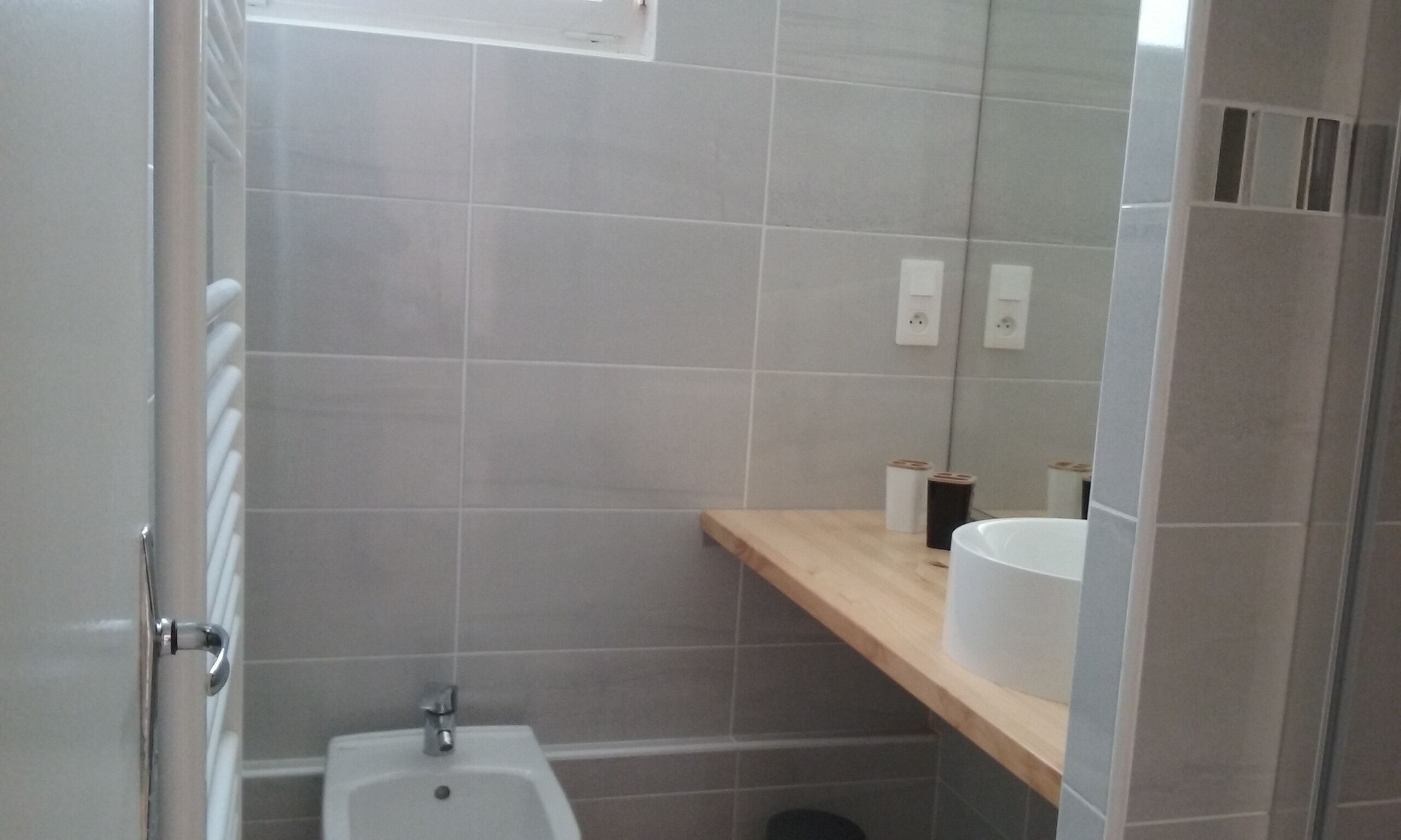 A bathroom with a bidet, a wooden shelf with a white container, and a mirror reflecting the room.