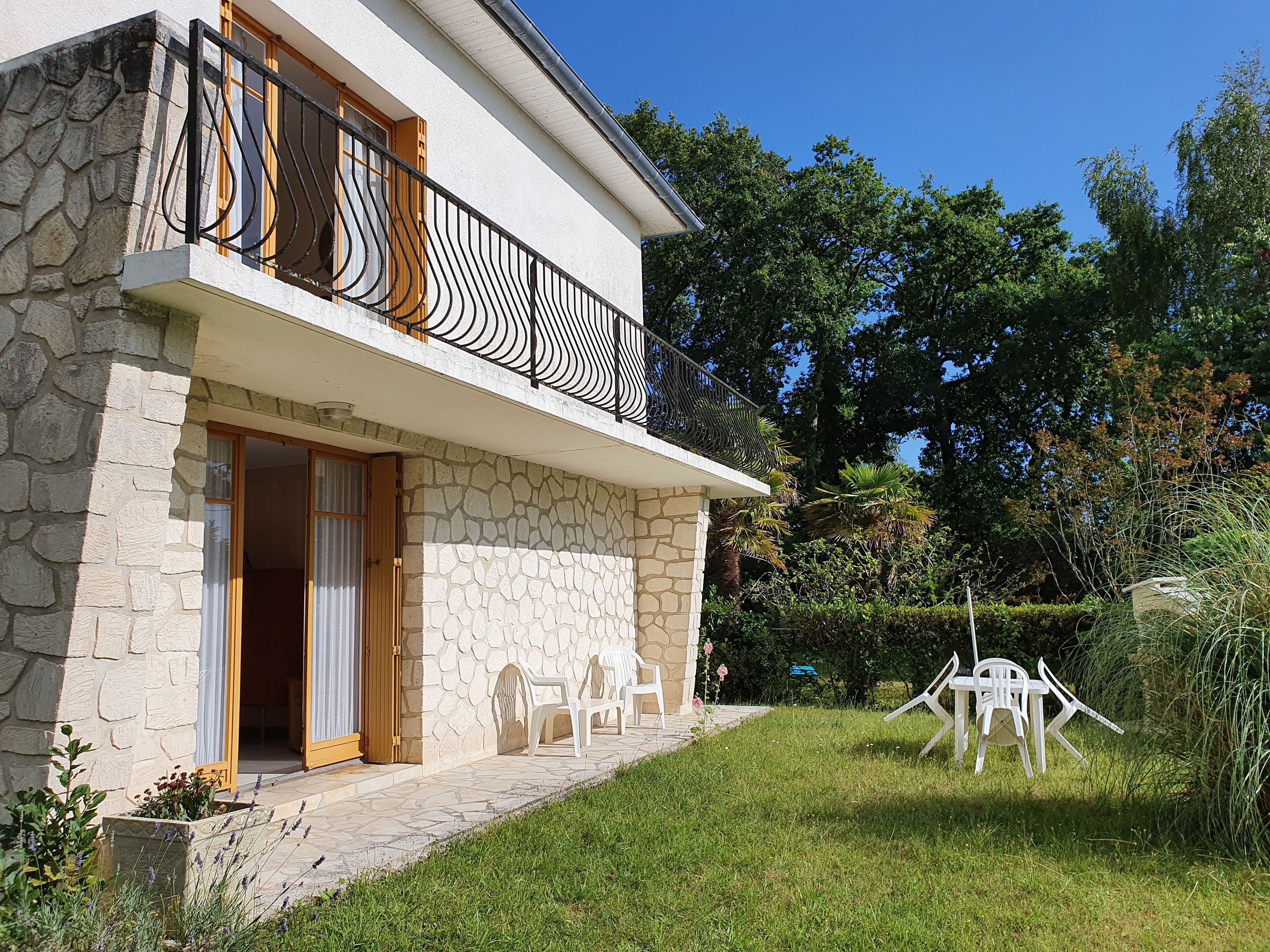 A two-story house with a stone facade, a balcony with black metal railings, and a small garden with plastic chairs.