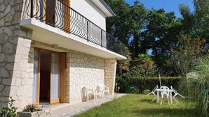 Ein zweigeschossiges Haus mit einer Stein-Fassade, einem Balkon mit schwarzen Metallgeländern und einem kleinen Garten mit Plastikstühlen.