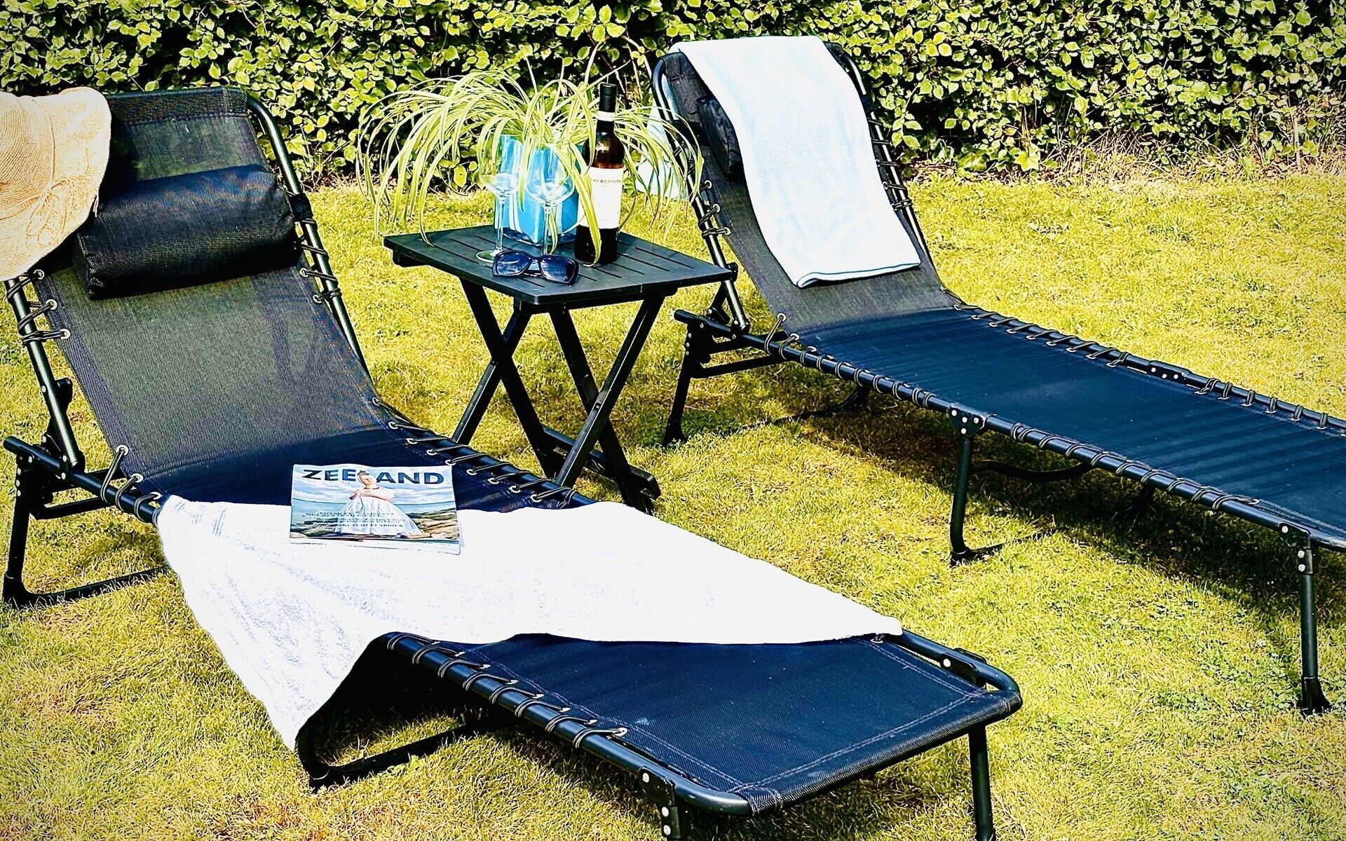 Two outdoor lounge chairs with a table and a bottle of wine between them, set on a grassy lawn with a hedge in the background.