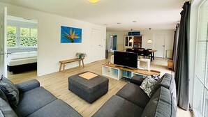A modern living room with a large gray sectional sofa, a wooden coffee table, and a flat-screen TV on a white stand. There is a painting of a blue flower on the wall, and a bench with a plant beside the door.