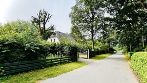 A narrow paved road flanked by lush greenery and a white building with a dark roof.