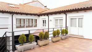 Un edificio con techo de tejas, paredes blancas y varias ventanas. En la planta baja, hay cinco jardineras rectangulares con árboles pequeños.