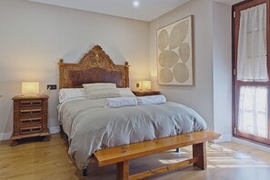 A bedroom with a wooden bed, bedside tables, a bench, a large abstract painting, and a window with curtains.