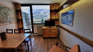 A room with a wooden dining table, chairs, a television on a wooden cabinet, a window with a view of mountains, and a poster on the wall.
