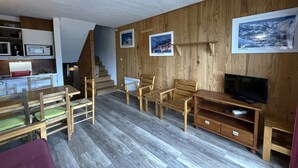 Un espai vital compacte amb un terra de fusta, una zona de menjador amb taula i cadires, una televisió sobre un suport de fusta i una escala que mena a un altre nivell.