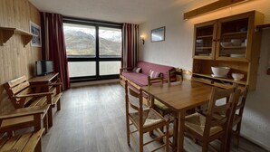 Habitació acollidora amb un banc de fusta, una taula de fusta i un sofà. Hi ha una finestra amb cortines, una televisió i una estant penjada a la paret amb plats.