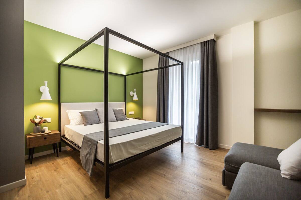 A modern bedroom with a four-poster bed, green walls, and a grey sofa.