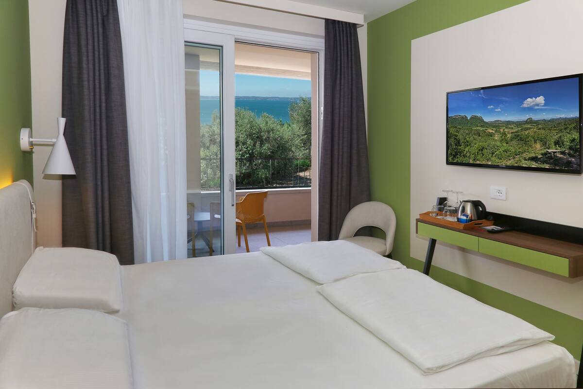 A hotel room with a large bed, a desk with a chair, a television, and a view of the sea through a sliding door.