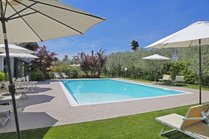 Et rektangulært pool med en grusramme, omgivet af en græsplæne med flere liggestole og parasoller.