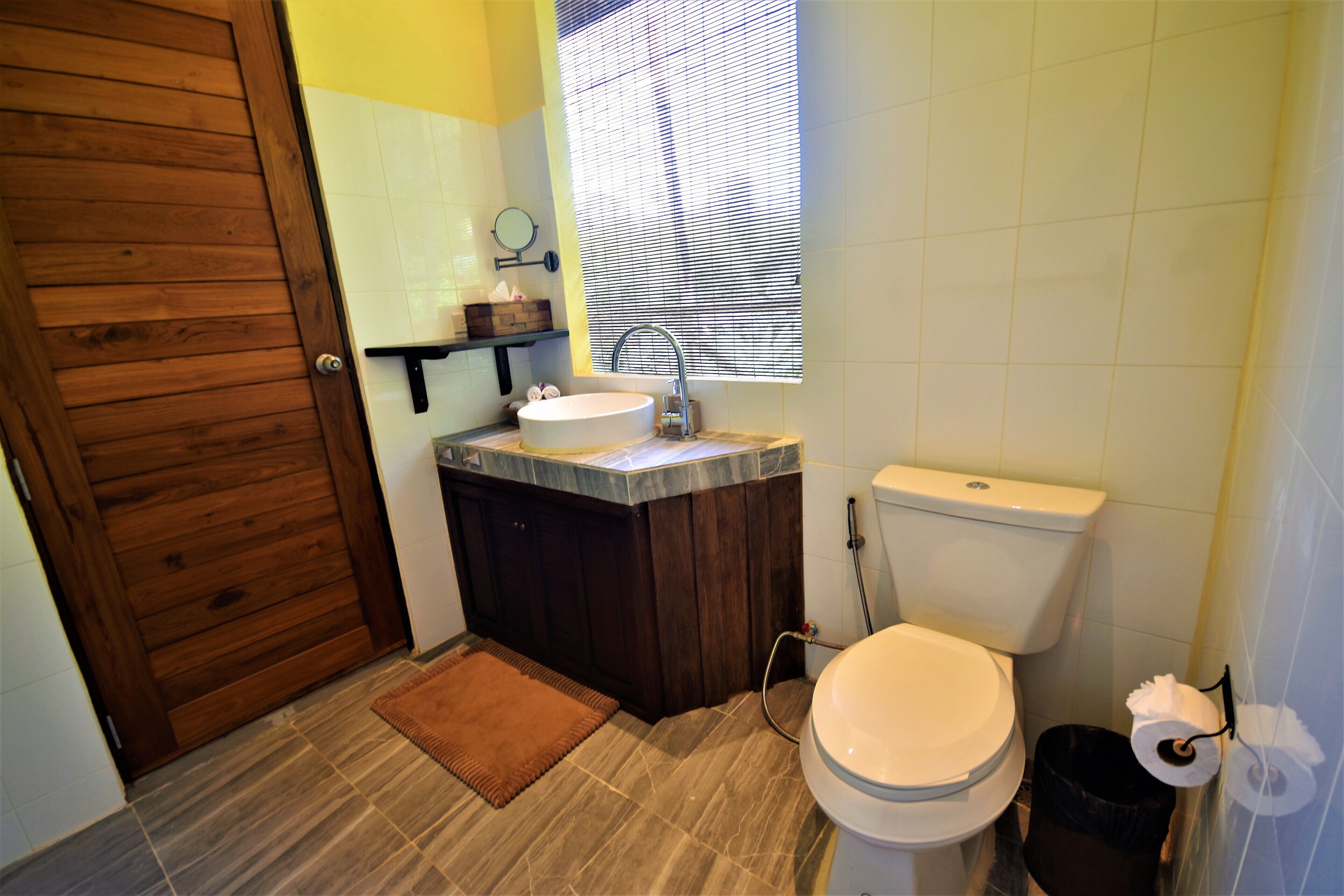 A bathroom with a wooden door, a sink, a toilet, and a window with blinds.