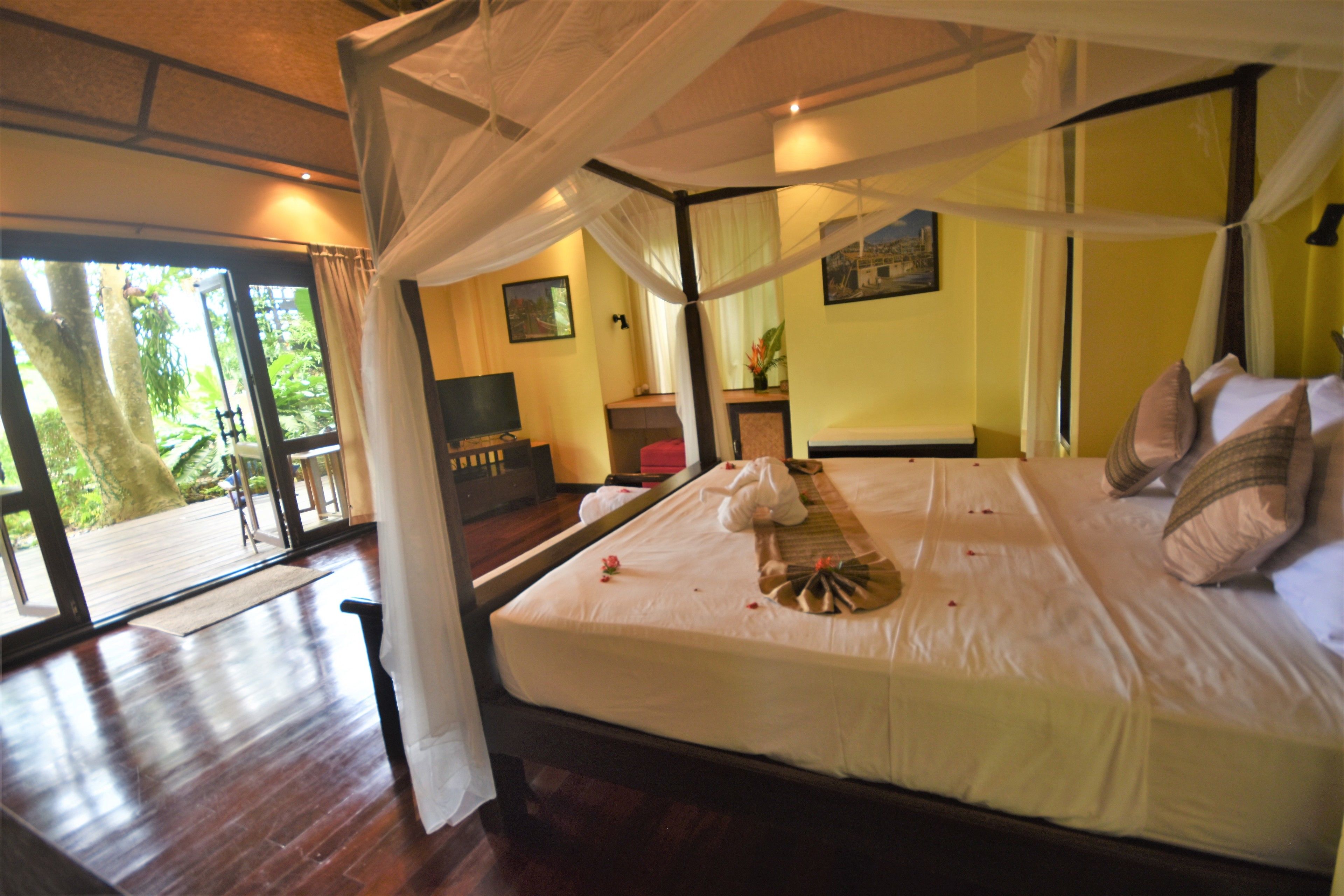 A four-poster bed with a canopy, a television, and a desk with a chair in a room with a large window.